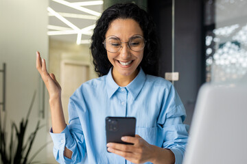 Wall Mural - Happy successful hispanic woman smiling reading notification, celebrating victory success, businesswoman received news online message, holding phone, using app on smartphone.