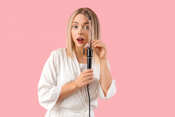 Wall Mural - Shocked young blonde woman curling hair on pink background