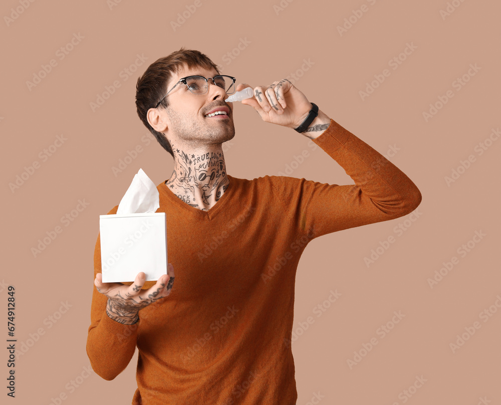 Allergic young man with tissues using nasal drops on brown background
