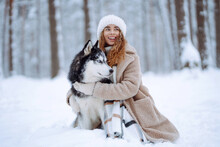 A Cheerful Husky Dog Walks With Its Owner In A Snowy Forest. A Young Woman With Her Pet On An Adventure. Friendship Concept, Pets.