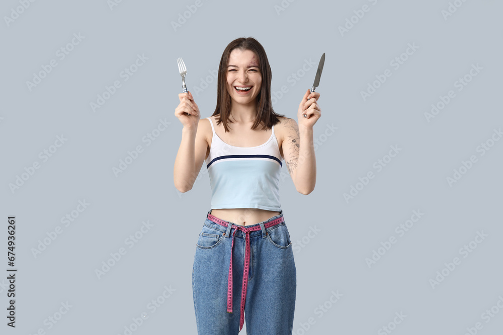 Beautiful young sporty woman with cutlery and measuring tape on grey background. Weight loss concept