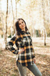 © Mila - A beautiful girl in jeans and a plaid shirt walks in a birch grove. Portraits of a brunette girl in nature, in the autumn forest