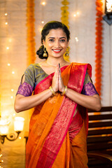 Vertical shot of happy indian girl with traditional ethnic dress showing namaste gesture by looking at camera during festival celebration at home - concept of welcome, greetings and Indian culture.