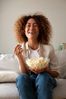 © Daniel - Vertical portrait of young multiracial hispanic woman laughing watching tv at home sitting on the sofa eating popcorn.