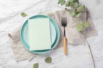 Wall Mural - Wedding menu card mockup on plate with linen napkin on marble table with green leaves