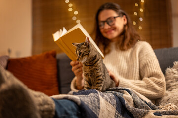 Сozy winter evening at home,woman sitting on the sofa with a gray cat in her arms reading a book