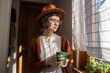 © DimaBerlin - Serious teenage girl in hat holding cup of tea standing at window and looking at camera. Teenager in big glasses with concentrated glance looks strange like retro girl. Sunlight shadow falling on face