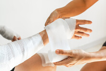 Sister Wrapping Her Brother Wrist And Arm With Bandage Around Injured Hand Over White Background At Home. First Aid, Accident And Injury Treatment Concept. Closeup
