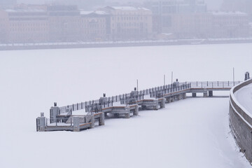  Pier on the city frozen river covered with snow. It's snowing over the city. Cold snowy winter.