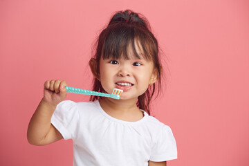 Cheerful little asian girl brushes teeth, has brilliant smile, morning daily hygiene, poses with toothbrush, isolated over pink background. Children dental health care concept.