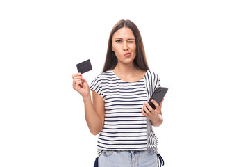 Wall Mural - young positive brunette lady with straight hair in glasses dressed in a striped t-shirt uses a credit card and a smartphone on a white background with copy space