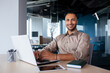 © Liubomir - Attractive african american man sitting at desk in front of laptop in office, programmer working in modern environment, tapping on keyboard and smiling at camera.