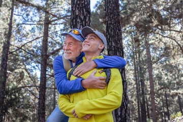 Wall Mural - Happy family couple of grandfather and young grandson hugging while hiking in the mountains expressing joy and freedom, healthy lifestyle together in the woods. Adventure is ageless