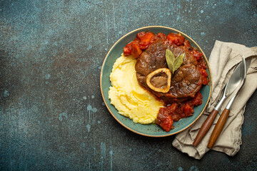 Wall Mural - Traditional Italian dish Ossobuco all Milanese made with cut veal shank meat with vegetable tomato sauce served with corn polenta on ceramic plate top view on rustic stone background, copy space.