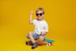 Adorable little boy in white tshirt and sunglasses, sitting on modern skateboard while looking at camera and pointing up, against yellow background.