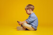 Side view of focused little boy sitting on studio floor with crossed legs while playing addictive games on smartphone, isolated over yellow background. Gadget addiction.