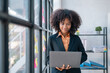 © amnaj - Young african american happy businesswoman standing and working on laptop by window in office.