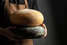 Craftsmanship Hands Have A Typical Italian Cheese. French Tomme Cheese In The Hands Of A Cheesemaker On Dark Background. Long Banner Format