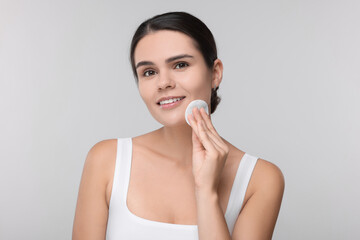 Poster - Young woman cleaning her face with cotton pad on light grey background