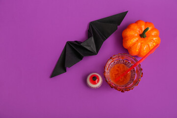 Wall Mural - Composition with glass of delicious cocktail for Halloween, candy, pumpkin and paper bat on purple background