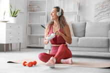 Smiling Adult Woman In Sportswear With Bottle Of Water Sitting On Yoga Mat At Home