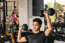 A Young Asian Man Wearing A Black Moisture-wicking Polyester Shirt Doing A Set Of Seated Alternate Dumbbell Presses At The Gym. Shoulder Workout And Training.