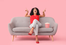 Beautiful Woman With Popcorn On Sofa Pointing At Something Against Pink Background