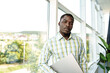 © fotofabrika - Young african man standing near the window indoors and holding laptop