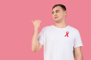 Sticker - Handsome young man with red ribbon pointing at something on pink background. World AIDS day concept