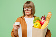 Elderly sad serious dissatisfied woman wearing brown shirt casual clothes glasses hold shopping paper bag with food products read check isolated on plain green background. Delivery service from shop.