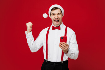 Wall Mural - Merry young man wear white shirt Santa hat posing hold use mobile cell phone do winner gesture isolated on plain red background studio portrait. Happy New Year Christmas celebration holiday concept.