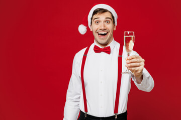 Wall Mural - Merry young surprised man wear white shirt Santa hat posing hold glass with champagne look camera isolated on plain red background studio portrait Happy New Year Christmas celebration holiday concept