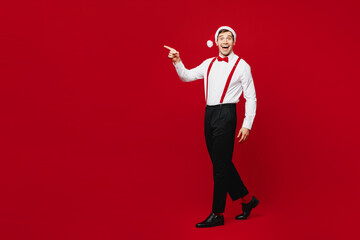 Full body merry young shocked fun man wear white shirt Santa hat posing walk go point index finger aside isolated on plain red background studio. Happy New Year Christmas celebration holiday concept.