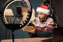Male influencer wearing Santa hat recording a video with his smartphone in a ring of light while unboxing a gift of VR Glasses.
