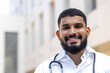 Close-up portrait of a young Indian male doctor standing outside a clinical center in a white coat and smiling at the camera
