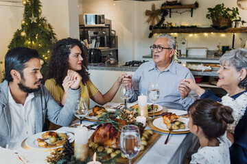 Wall Mural - Latin Family praying and holding hands for prayer gathering at thanksgiving dinner at home in Mexico. Grateful people with gratitude enjoy a turkey meal in Latin America