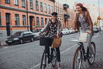 Wall Mural - Two stylish young female friends biking in the city