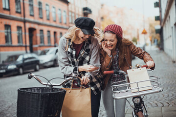 Wall Mural - Two stylish young female friends biking in the city