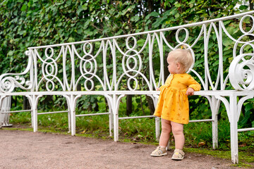 A cute little baby girl with a ponytail on her head is standing by a beautiful park bench