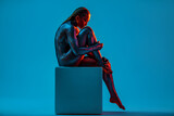 Nude woman with black gloss paint on body sitting on cube on blue background