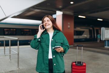 Wall Mural - Redhead woman waiting train and using smart phone. Railroad transport concept, Traveler. 30s Middle age woman with red suitcase and passport talking on smartphone, walking at railroad station platform