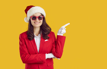 Christmas banner with smiling brunette woman in Santa hat, sunglasses, white gloves and red suit pointing on copy space on yellow background. New Year, celebrating winter holidays, Xmas advertisement.