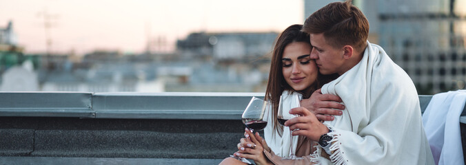 Beautiful young loving couple on a surprise romantic date on a roof top. Picturesque view, skyscrapers on background. Wine, candles, kisses on holiday Saint Valentine's Day celebration outdoors