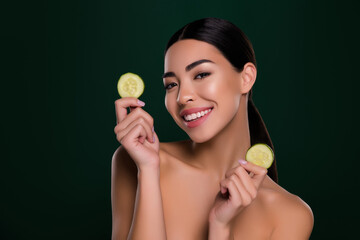 Wall Mural - Photo of gorgeous asian young lady hold cucumber slices spa salon shoulders off isolated dark green color background