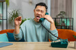 Tired bored Asian man talking on wired vintage telephone of 80s, fooling, making silly faces, exhausted of tedious story, not interested in communication talk at home room. Guy sits on couch at table