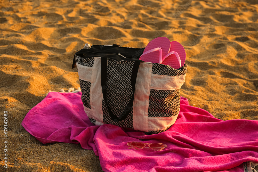 Stylish beach bag with flip-flops, sunglasses and towel on sand