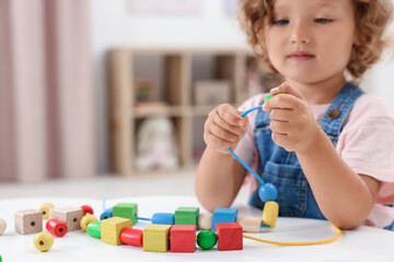 Wall Mural - Motor skills development. Little girl playing with wooden pieces and string for threading activity at table indoors, closeup