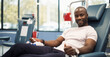 Black Man Donating Blood For People In Need In Bright Hospital. African Male Donor Squeezing fist To Pump Blood Through The Tubing Into The Bag. Donation for Children Battling Cancer.