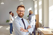 © Studio Romantic - Portrait of happy smiling confident bearded business man leader in glasses proudly looking at the camera with crossed arms and with team of company employees talking in background in the office.