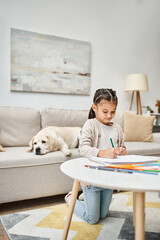 Wall Mural - little girl in casual clothes drawing on paper with color pencil near labrador in living room, art